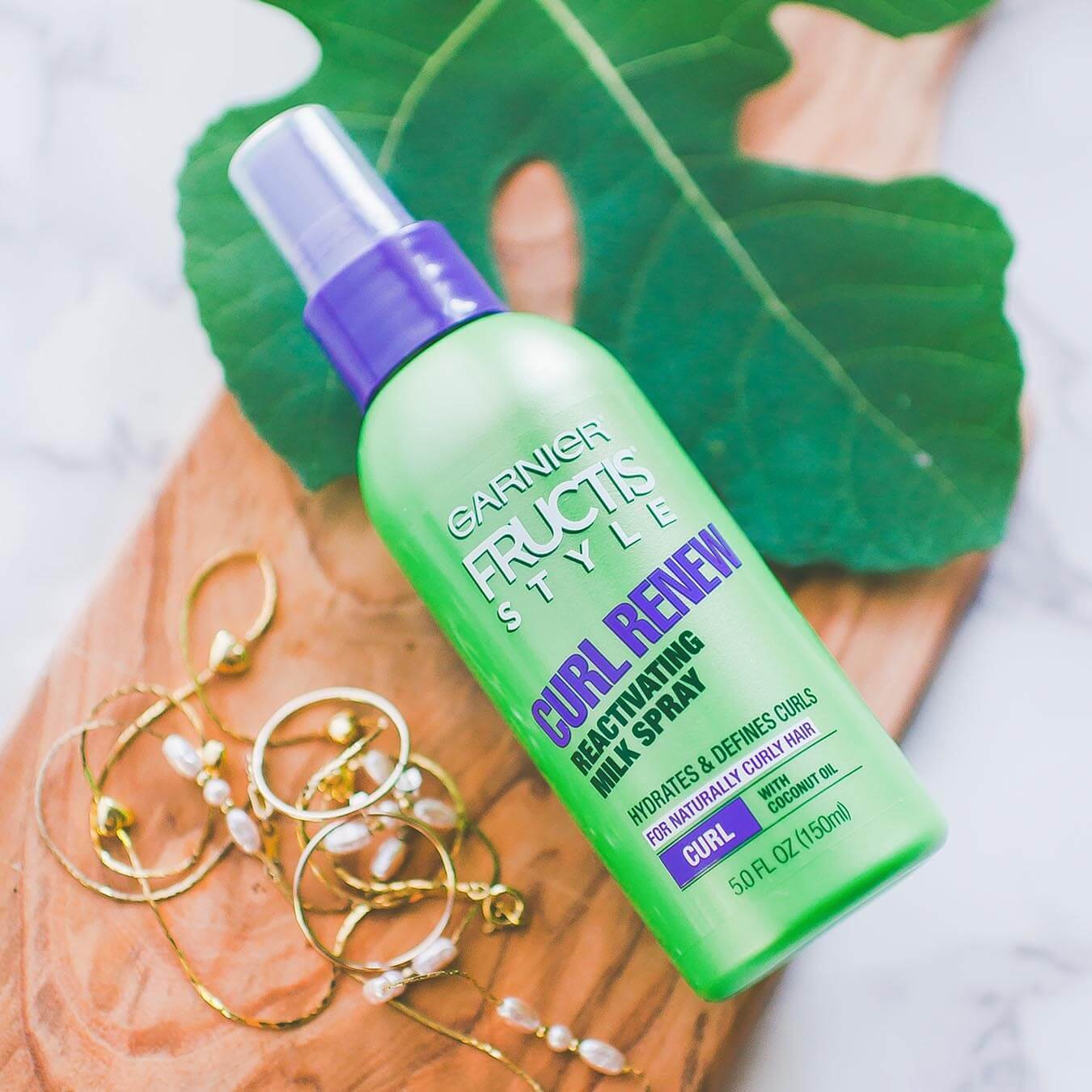 Garnier Fructis Style Curl Renew Reactiving Milk Spray on a wooden oar next to a large sassafras leaf and gold jewelry.